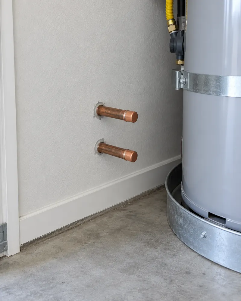 Two capped copper pipes stubbed out of a garage wall near the floor beside a water heater, the classic pre-plumbed water softener loop